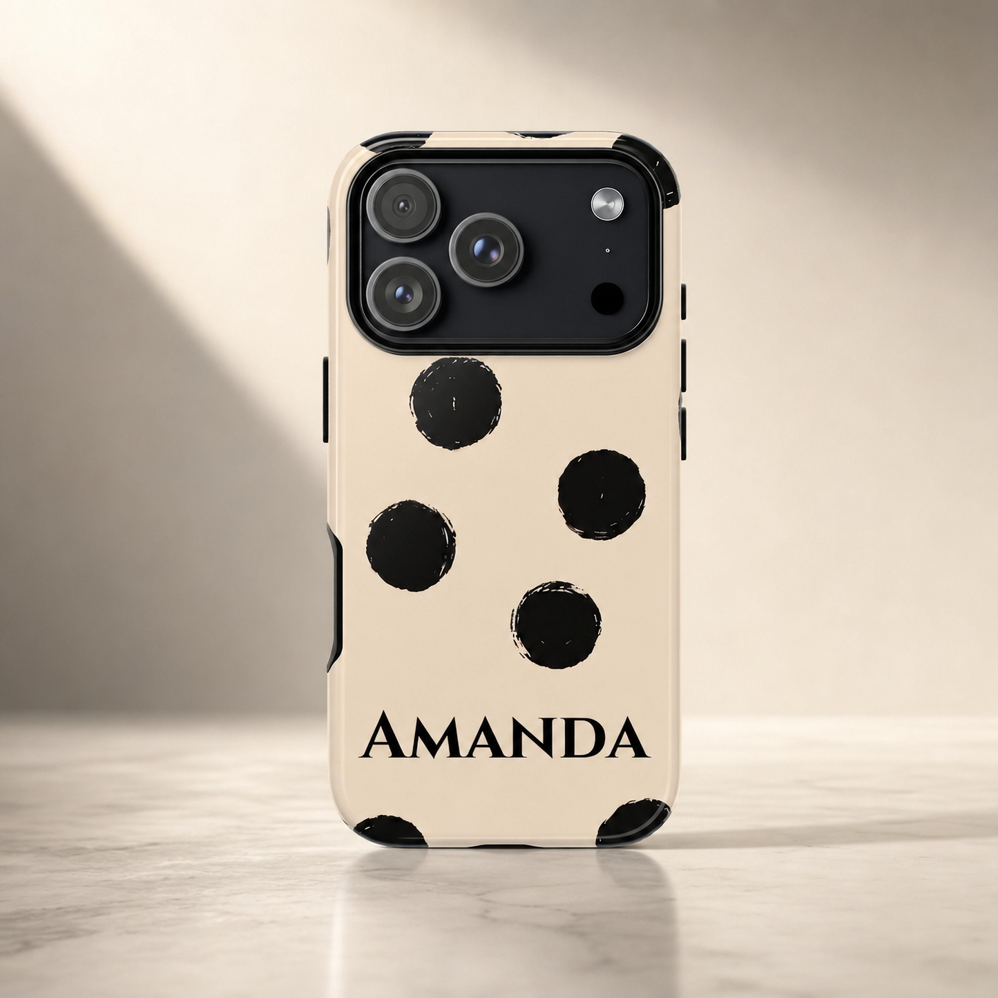 A cream-colored phone case with black polka dots and the name 'AMANDA' printed in black at the bottom, shown on a smartphone standing upright on a light marble surface against a warm beige background.