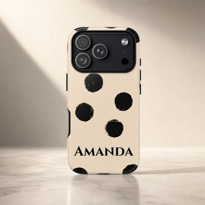 A cream-colored phone case with black polka dots and the name 'AMANDA' printed in black at the bottom, shown on a smartphone standing upright on a light marble surface against a warm beige background.