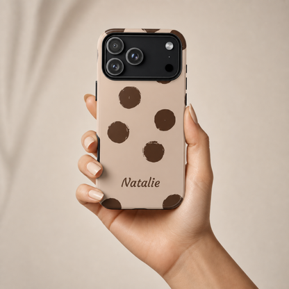 A hand holding a beige phone case with dark brown polka dots and the name 'Natalie' printed near the bottom. The phone case is shown against a soft, neutral beige background.