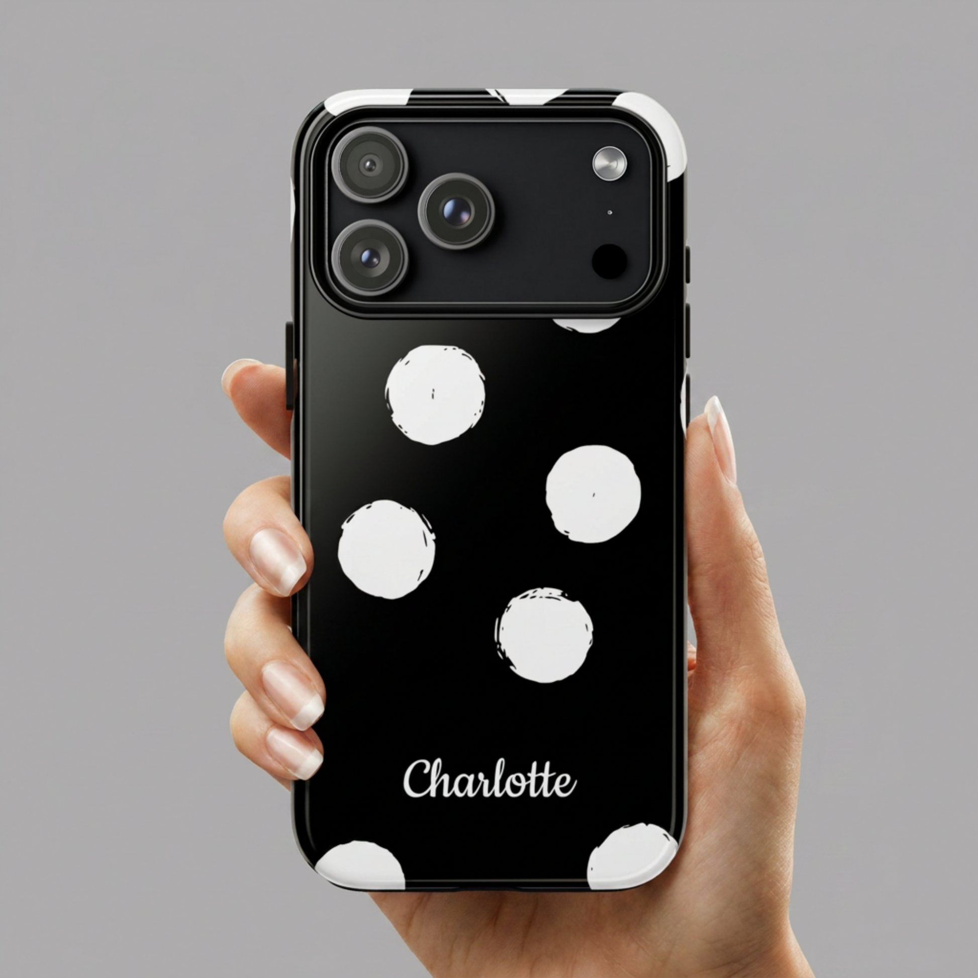 A hand holding a black phone case with white polka dots and the name 'Charlotte' printed in white script near the bottom, shown against a plain gray background.