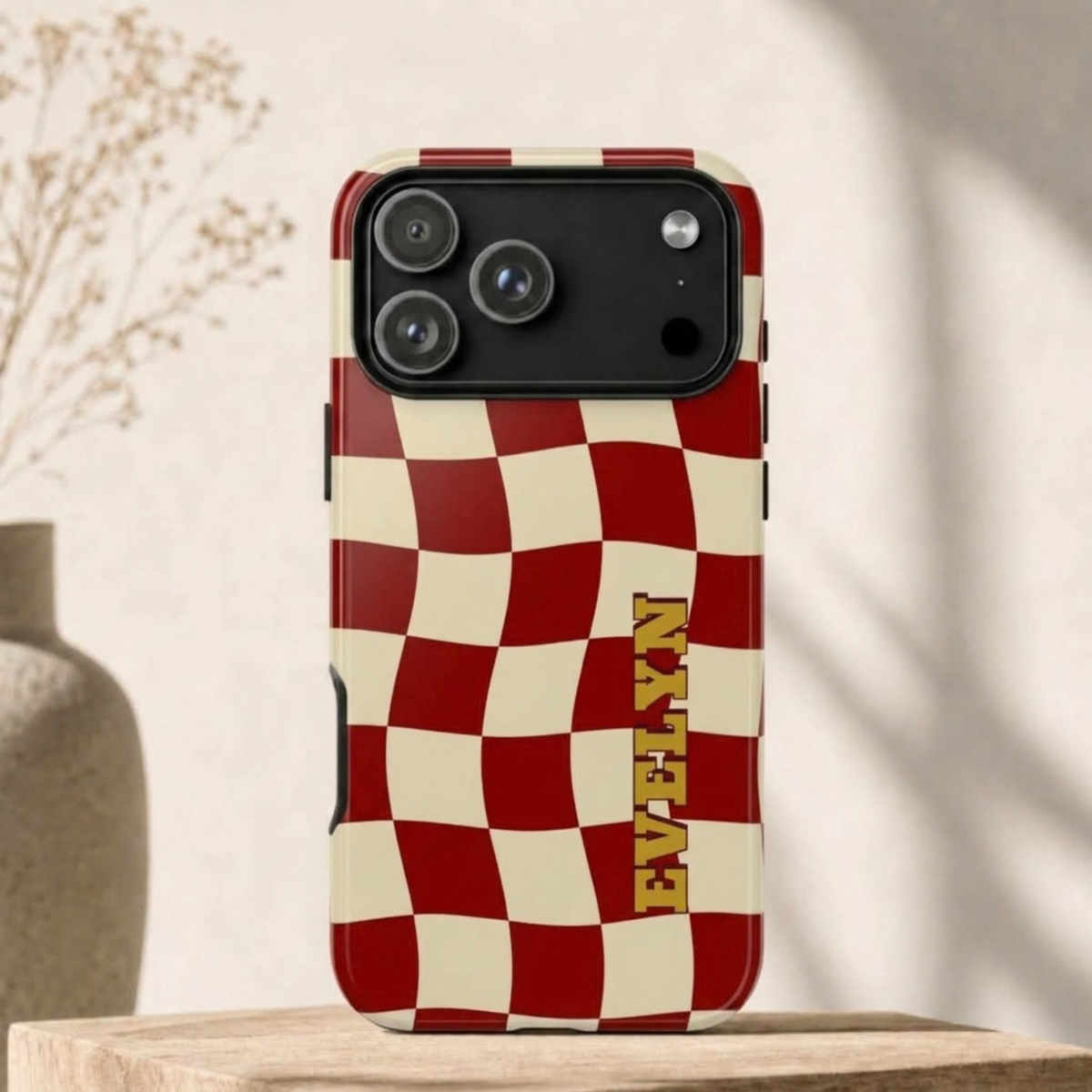 A phone case with a red and cream checkered pattern and the name 'EVELYN' printed vertically in gold, shown standing on a wooden surface against a softly lit beige background.