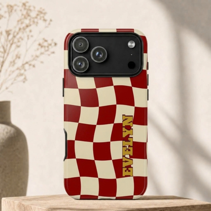 A phone case with a red and cream checkered pattern and the name 'EVELYN' printed vertically in gold, shown standing on a wooden surface against a softly lit beige background.
