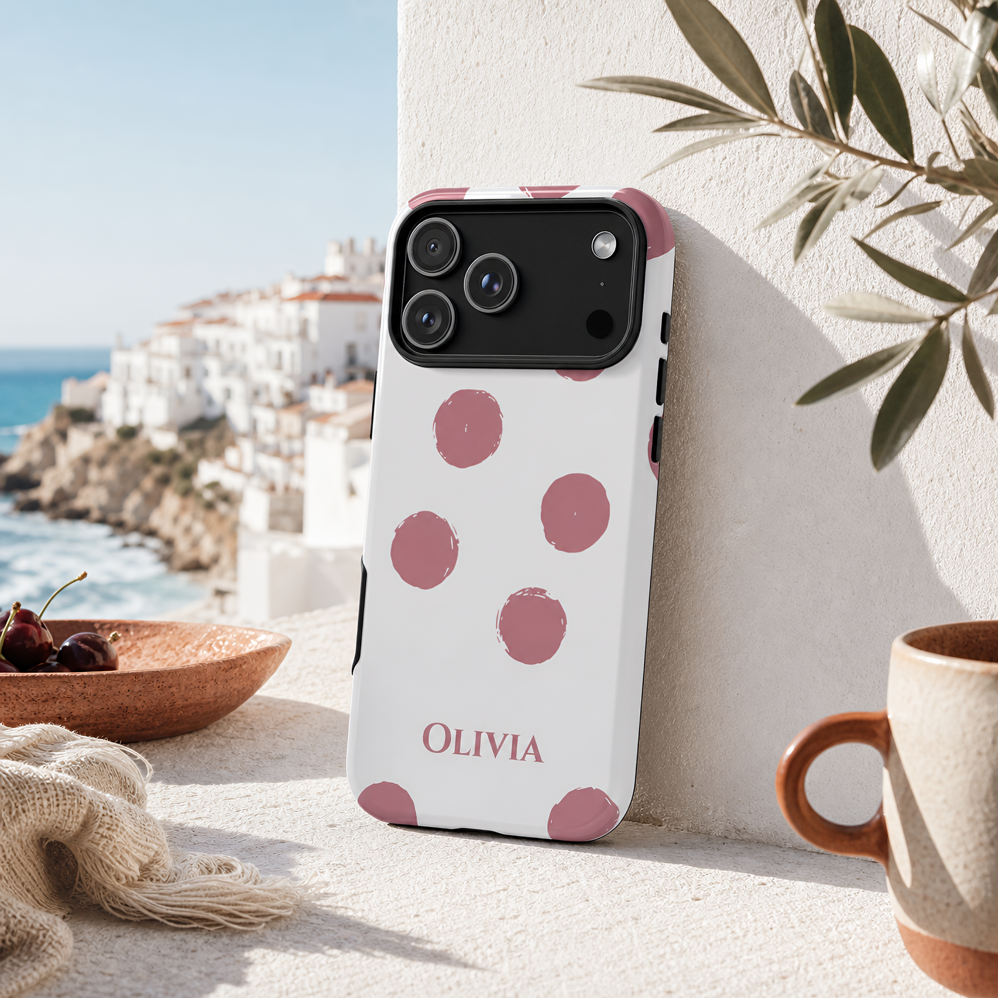 A white phone case with pink polka dots and the name 'OLIVIA' printed in pink at the bottom, shown leaning against a white wall in a sunlit outdoor setting with a coastal background.